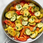 Vibrant sautéed vegetables, including yellow squash, zucchini, carrots, and red bell peppers, seasoned with herbs in a pan.