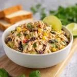 A vibrant bowl of southwest chicken salad with corn, black beans, and cilantro, served with lime wedges and bread.