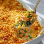 Creamy vegetable casserole with a golden, crispy breadcrumb topping, being served from a white baking dish, showing broccoli, corn, peas, and carrots.