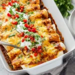 A close-up of a white baking dish filled with cheesy Beef and Bean Enchiladas casserole topped with sour cream, tomatoes, and fresh cilantro.