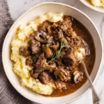 A close-up shot of a bowl filled with creamy mashed potatoes generously topped with savory beef tips and gravy, garnished with fresh rosemary.