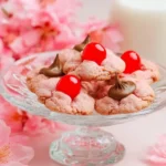 Pink Cherry Blossom Cookies topped with maraschino cherries and chocolate kisses on a glass cake stand with a blurred background of pink flowers and milk.