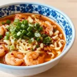 A vibrant bowl of Chinese Shrimp Noodle Soup garnished with fresh green onions and red chili flakes on a light wooden table.