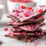 A delightful stack of pink and dark chocolate bark topped with red heart candies and colorful sprinkles, perfect for a sweet treat.