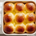 A white baking dish filled with nine glistening golden-brown Copycat Texas Roadhouse Rolls, sitting in melted butter.