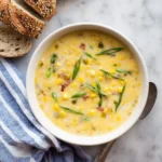 A bowl of creamy corn soup garnished with green onions and bacon bits, served with sliced rustic bread on a marble background.