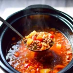 A steaming ladle full of hearty Crockpot Hamburger Soup with ground beef, potatoes, carrots, and bell peppers, being served from a slow cooker.