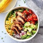 A beautiful grilled chicken salad bowl with sliced chicken, corn, cherry tomatoes, avocado, red onion, and crumbled cheese.