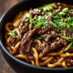 A close-up shot of a black bowl filled with delicious Hoisin Beef Noodles, garnished with sesame seeds and chopped green onions.