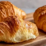 Three golden homemade French croissants on a wooden board, with one broken open revealing its flaky, airy interior.