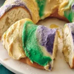 A vibrant lemon king cake, beautifully decorated with purple, green, and gold icing and sprinkles, with a slice removed showing its soft interior.