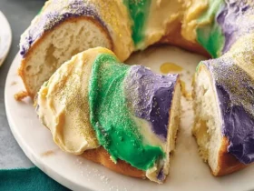 A vibrant lemon king cake, beautifully decorated with purple, green, and gold icing and sprinkles, with a slice removed showing its soft interior.