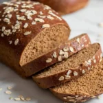 A freshly baked loaf of Molasses Brown Bread, sliced to reveal its rich texture and topped with oats, on a white surface.