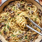 A close-up shot of a creamy Mushroom and Garlic Spaghetti dish being served from an orange pot with tongs, garnished with fresh parsley.