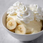 A white bowl filled with creamy Ninja Creami Banana Cream Pie Ice Cream, topped with swirls of whipped cream and fresh banana slices.