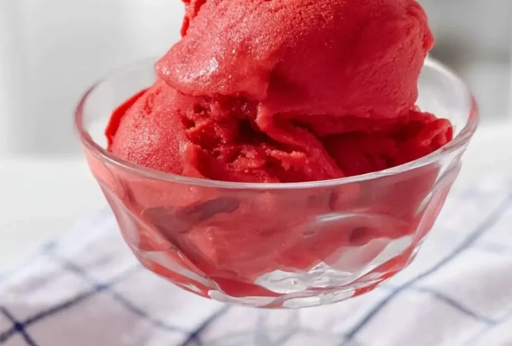 A clear glass dish holds several scoops of vibrant red Ninja Creami Strawberry Sorbet on a checkered cloth.