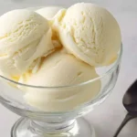A clear glass dessert bowl filled with multiple scoops of creamy Ninja Creami Vanilla Ice Cream, with a silver spoon nearby.