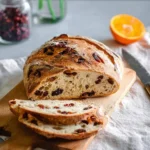 A rustic loaf of Orange Cranberry Sourdough Bread, partially sliced on a wooden cutting board, with an orange half and a jar of cranberries in the background.