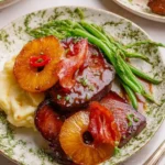 A visually appealing plate featuring Pineapple Brown Sugar Glazed Beef, topped with grilled pineapple and bacon, served with mashed potatoes and asparagus.