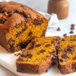 A freshly baked loaf of Pumpkin Chocolate Chip Bread, golden brown with visible chocolate chips, partially sliced on a white cutting board.