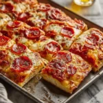 A delicious, golden-brown pepperoni Sheet Pan Pizza, cut into square slices on a baking sheet, with a glass of beer in the background.