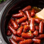 Close-up of delicious Slow Cooker Mississippi Lil Smokies simmering in a rich, glossy sauce in a black crock pot with a wooden spoon.