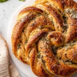 A golden-brown Sourdough Herb Garlic Twist Loaf, beautifully braided and topped with herbs and cheese, rests on a white marble surface.