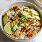 A delicious street corn chicken rice bowl with seasoned chicken, corn, avocado, jalapeños, creamy sauce, and a fresh lime wedge.
