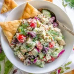 A fresh tzatziki chicken salad with chicken, tomatoes, olives, cucumber, and dill, served in a white bowl with pita triangles.