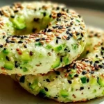 Close-up of two vibrant green Zucchini Bagels topped with a generous sprinkle of black and white sesame seeds on a plate.