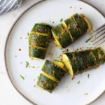 Four seasoned Air Fryer Hasselback Zucchini, sliced and ready to eat, are artfully arranged on a white plate with a fork.