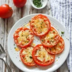 A white plate full of delicious air fryer parmesan tomatoes, garnished with melted cheese and fresh herbs, on a rustic wooden table.
