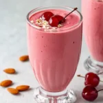 A vibrant pink almond cherry smoothie in a decorative glass, garnished with two fresh cherries and oats, surrounded by whole almonds.