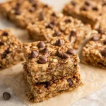 A close-up of a stack of three chocolate chip banana oatmeal bars on parchment paper, with more blurred bars in the background.