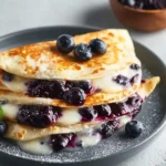 A stack of golden-brown Blueberry Breakfast Quesadillas filled with creamy sauce and blueberry compote, topped with fresh blueberries and powdered sugar on a grey plate.