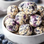 White bowl of Blueberry Cheesecake Protein Bites, showing oats, chia seeds, and blueberry swirls. Fresh blueberries are scattered nearby.