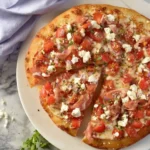 A top-down view of a freshly baked Bruschetta Pizza, generously topped with diced tomatoes, feta cheese, prosciutto, and herbs, served on a white plate.