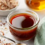 A clear glass jar of Carolina Vinegar BBQ Sauce, a spoon with a drop of sauce, and scattered red chili flakes on a white surface.