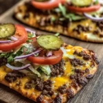 Two delicious Cheeseburger Flatbreads topped with ground beef, melted cheese, lettuce, tomato, onion, and pickles on a wooden board.