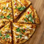 A close-up overhead shot of a delicious Chicken Enchilada Pizza on a wooden board, garnished with corn, peppers, cilantro, and a creamy drizzle.