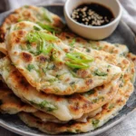 A close-up of a stack of golden-brown Chinese Scallion Pancakes garnished with fresh scallions, with a dipping sauce.