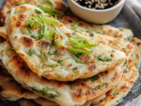 A close-up of a stack of golden-brown Chinese Scallion Pancakes garnished with fresh scallions, with a dipping sauce.