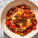 A beautifully plated bowl of pan-seared Cod with Tomatoes, green olives, and herbs over pasta, resting on a marble surface.