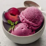 A white bowl holds three scoops of vibrant purple Creamy Cardamom Plum Sorbet, garnished with plum halves and a fresh green herb sprig.