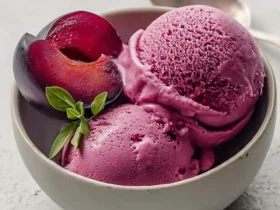 A white bowl holds three scoops of vibrant purple Creamy Cardamom Plum Sorbet, garnished with plum halves and a fresh green herb sprig.