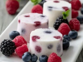 Several pink and white frozen yogurt berry bites are artfully arranged on a white plate, garnished with fresh mixed berries.