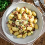 A white bowl filled with creamy Gnocchi Carbonara, topped with crispy bacon, grated cheese, and fresh parsley, on a rustic wooden table.