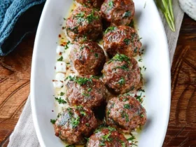 A white oval platter holds nine delicious Greek Beef Meatballs garnished with fresh parsley and a creamy sauce, set on a wooden table.