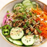 A vibrant and colorful Healthy Banh Mi Bowl, featuring seasoned ground meat, sliced cucumbers, pickled red onions, shredded carrots, jalapeños, and a creamy drizzle, served over white rice.
