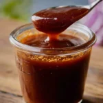 A spoon lifts thick, glossy homemade BBQ sauce from a glass jar, revealing its rich texture and deep reddish-brown color.